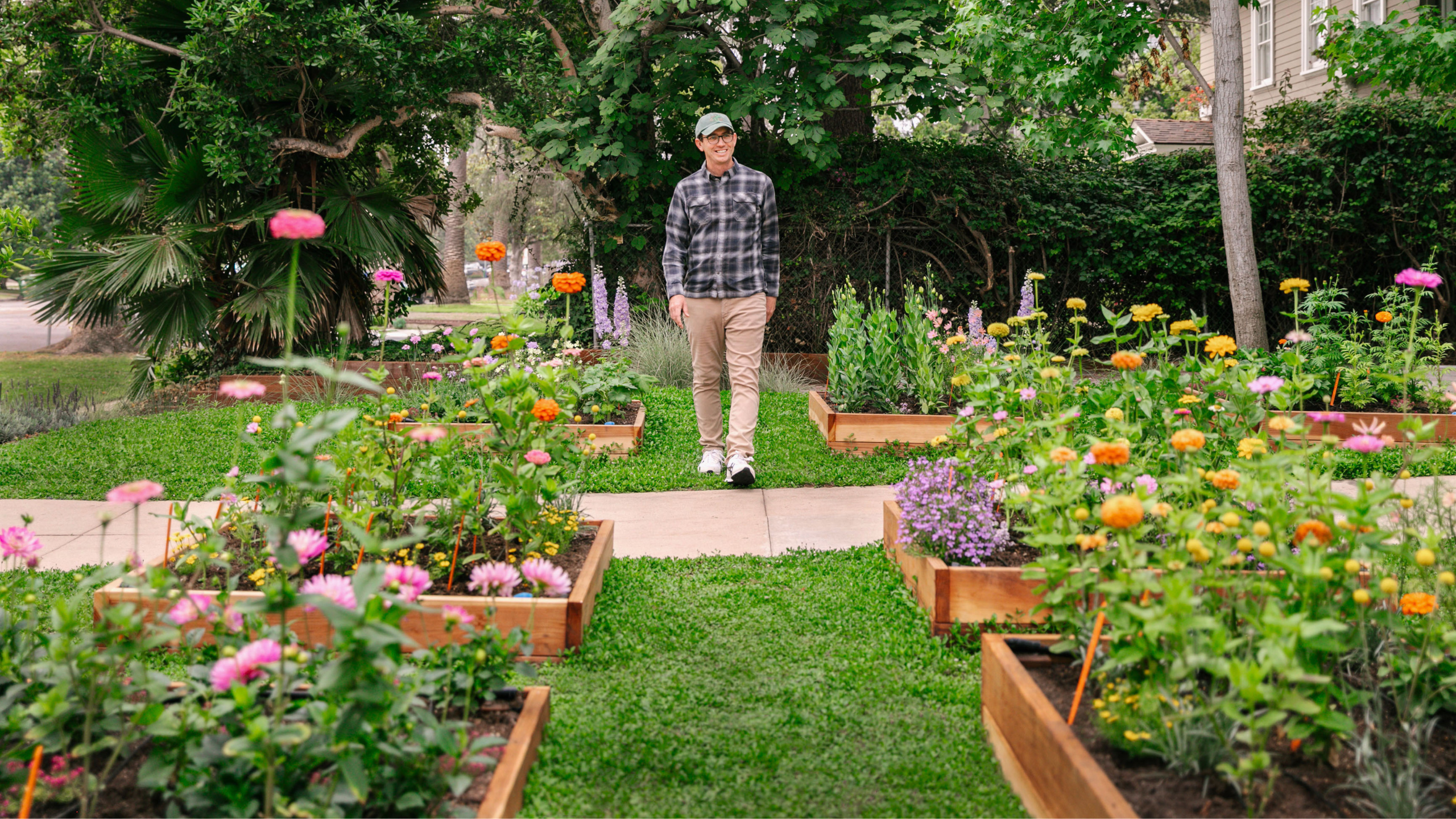 Person walking along a garden path surrounded by raised beds overflowing with colorful pink, orange, yellow, and purple flowers.