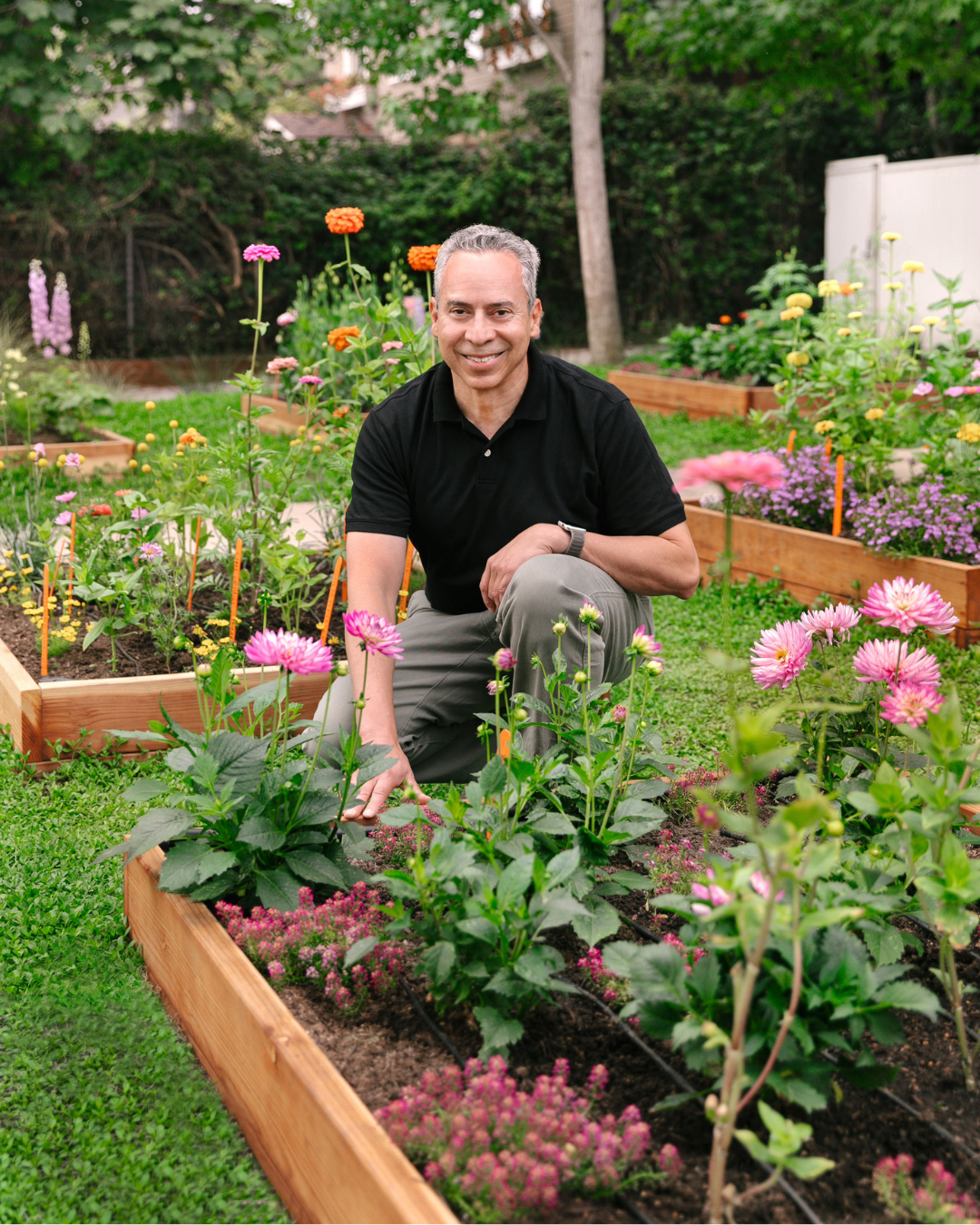 Landscape designer kneeling beside a wooden raised garden bed filled with pink flowers, surrounded by colorful blooms and lush greenery.